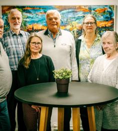 Et billede af seniorrådet. Syv personer står rundt om et bord og smiler. 