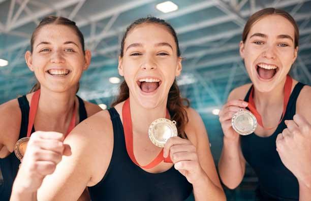 Tre unge kvinder i svømmedragter jubler og smiler, mens de holder en medalje frem foran brystet.