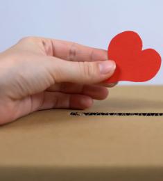 En hånd holder et lille udklippet hjerte foran en  papkasse, hvor der er skåret en revne i.