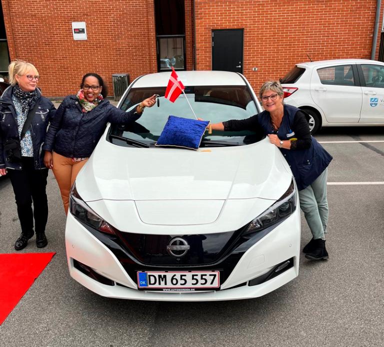 Tre kvindelige medarbejdere står ved en hvid mindre personbil og holder et flag og en bilnøgle frem henover kølerhjelmen