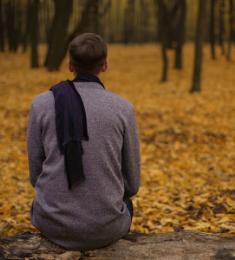 Yngre mand sidder med et halstørklæde, der er slynget om på ryggen, i en skov, hvor skovbunden er dækket af gult efterårsløv.