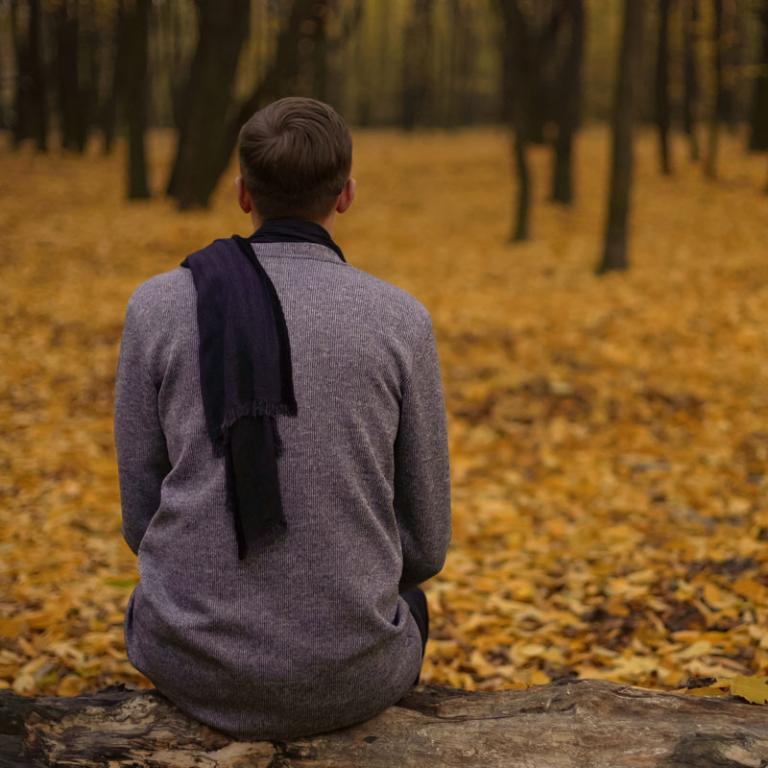 Yngre mand sidder med et halstørklæde, der er slynget om på ryggen, i en skov, hvor skovbunden er dækket af gult efterårsløv.