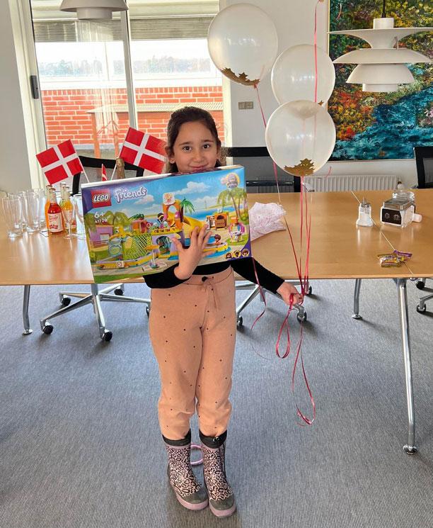 Pige holder med en pakke lego foran sig med den ene arm og i den anden har hun fire balloner med stjerner indeni. Der er flag på bordet i baggrunden.
