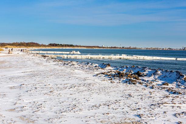 Kyststrækning med sneklædt strand med en bådebro, hvor der hænger is på. I baggrunden blå himmel.