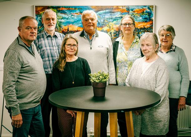 Et billede af seniorrådet. Syv personer står rundt om et bord og smiler. 