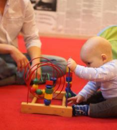 En baby sidder og leger med et stativ med klodser. En far i skjorte sidder på gulvet ved siden af.