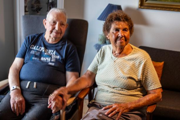 En ældre herre og dame sidder i hver sin stol og holder hinanden i hænderne.