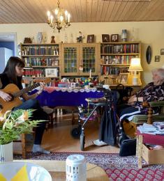 Ung kvinde sidder med en guitar og spiller foran en ældre kvinde i en gammel spisebordsstol.
