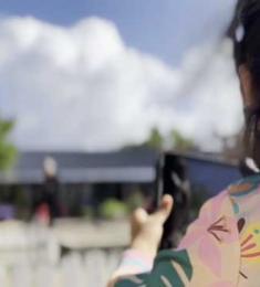 Pige med tablet står ved en legeplads og holder tabletten op for at tage foto. I baggrunden er himlen blå med hvide skyer