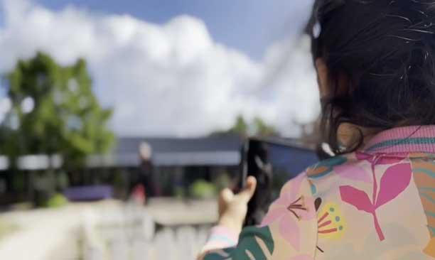Pige med tablet står ved en legeplads og holder tabletten op for at tage foto. I baggrunden er himlen blå med hvide skyer
