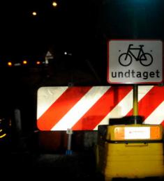 Mørk vejbane spærret af bom og skilt om undtagelse for cyklister. Skiltene oplyses af lyset fra forlygterne af en bil