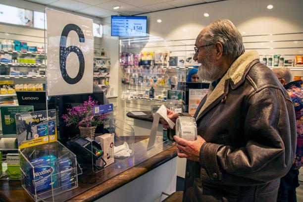 Ældre mand står ved skranken på et apotek med en bon og en rulle plastik i hånden.