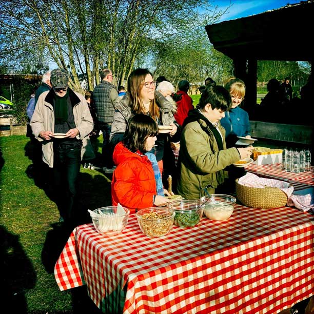 En kvinde og nogle børn står under åben himmel forrest ved et rødt og hvidternet bord dækket med mad. Bag dem en større folkemængde.