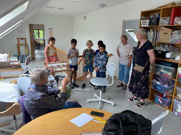 En gruppe af børn står i et loftsrum og kigger på en ældre kvinder, der har lavet håndarbejde.