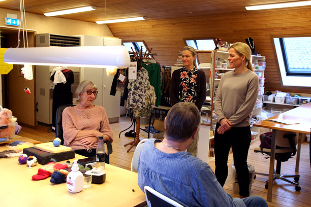 To ældre kvinder sidder med filt og stoffer og laver håndarbejde, mens ældreministeren og leder af seniorhuset Korsagergaard taler med dem.