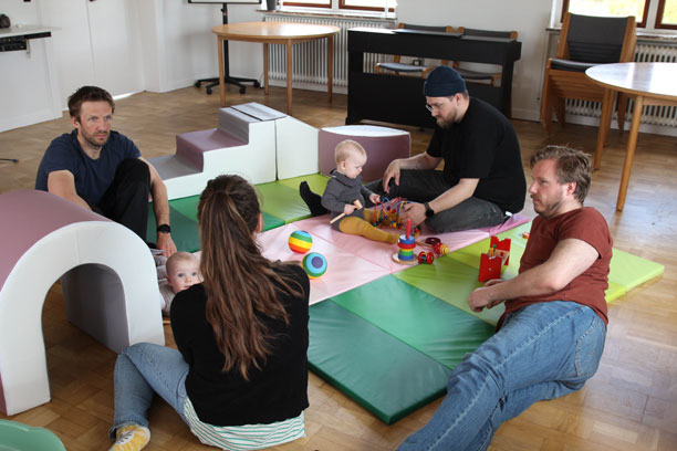 Mænd og babyer sidder på gulvet sammen med en kvindelig sundhedsperson