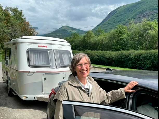 En kvinde står med armen på en bildør. Bag bilen en campingvogn og i baggrunden nogle grønne højdedrag