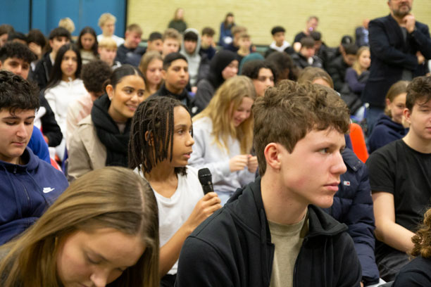 En ung mand med fletninger taler i en mikrofon. Han sidder i et publikum fyldt med unge mennesker. 